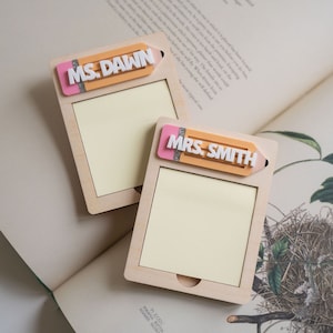 Puede incluir: Dos blocs de notas de madera con un diseño de lápiz en la parte delantera. El lápiz es rosa y amarillo con purpurina. Los blocs de notas están etiquetados como "MS. DAWN" y "MRS. SMITH".