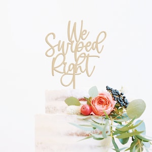 May include: Wooden cake topper with the text "We Swiped Right" in a cursive font. The topper is on a white cake with pink and green flowers.
