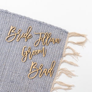 Peut inclure: Des panneaux en bois avec les mots "Bride Jillian" et "Groom Brad" sur une surface texturée grise avec des franges.