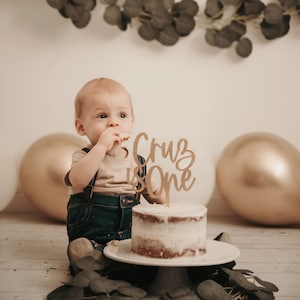Puede incluir: Un beb&eacute; con un peto vaquero y una camiseta color beige est&aacute; sentado cerca de un pastel peque&ntilde;o con un adorno de pastel que dice "Cruz is One". El pastel est&aacute; sobre un soporte blanco, con globos dorados y blancos en el fondo.