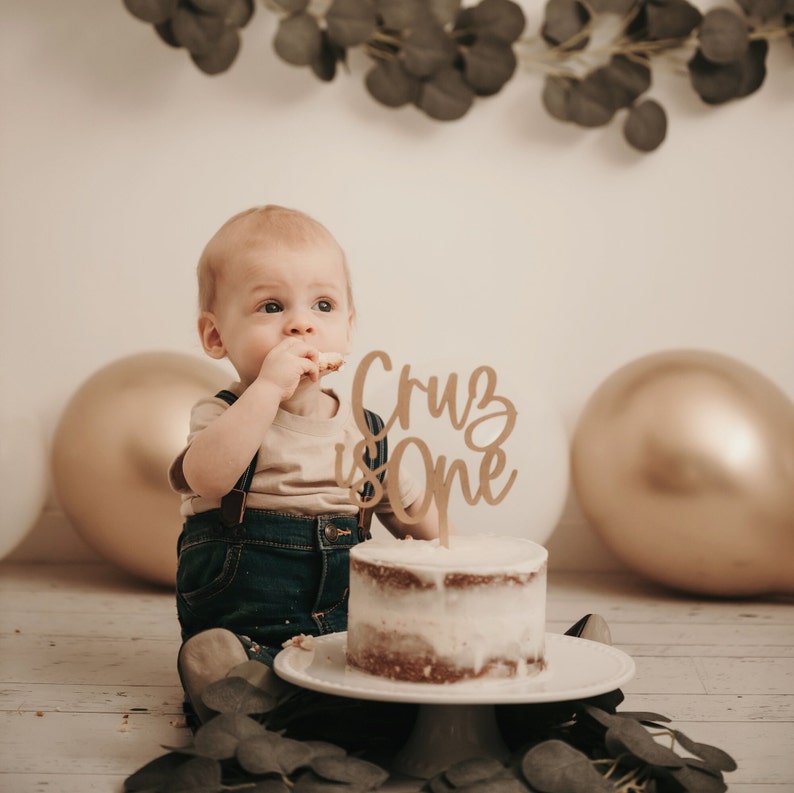 Puede incluir: Un beb&eacute; con un peto vaquero y una camiseta color beige est&aacute; sentado cerca de un pastel peque&ntilde;o con un adorno de pastel que dice "Cruz is One". El pastel est&aacute; sobre un soporte blanco, con globos dorados y blancos en el fondo.