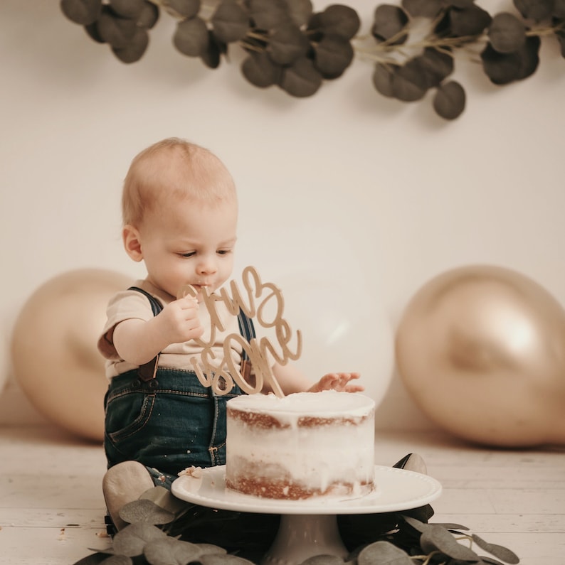 Puede incluir: Un pastel peque&ntilde;o y redondo con glaseado blanco sobre un soporte blanco para pasteles. Un adorno de pastel de madera dorada que dice "It's One". El pastel est&aacute; frente a un beb&eacute; con un peto de mezclilla. Globos dorados y blancos en el fondo.