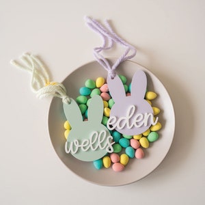 Puede incluir: Dos etiquetas de regalo en forma de conejo pastel con los nombres "Eden" y "Wells" escritos en ellas. Las etiquetas est&aacute;n rodeadas de huevos de caramelo de colores pastel en un plato gris claro.
