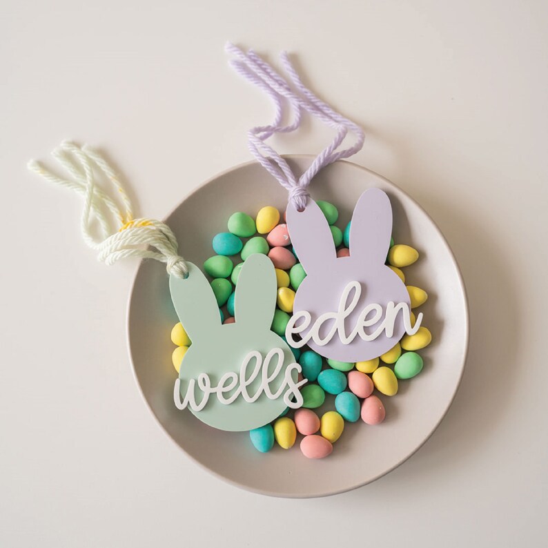 Puede incluir: Dos etiquetas de regalo en forma de conejo pastel con los nombres "Eden" y "Wells" escritos en ellas. Las etiquetas est&aacute;n rodeadas de huevos de caramelo de colores pastel en un plato gris claro.