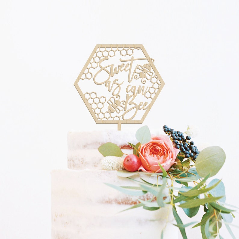 May include: Wooden cake topper in the shape of a hexagon with a honeycomb pattern and the text "Sweet as can Bee".