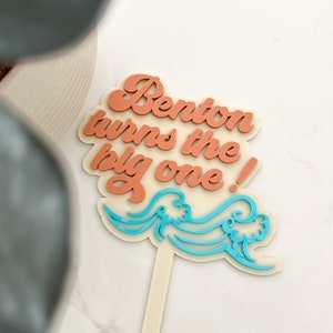 May include: A cake topper with the text "Benton turns the big one!" in peach-colored script with blue wave accents.