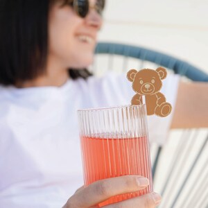 Puede incluir: Un vaso transparente y acanalado lleno de una bebida rosa, sostenido por una persona que lleva una camiseta blanca. Un agitador de bebidas con forma de oso de peluche marrón está unido al vaso. El fondo está borroso.
