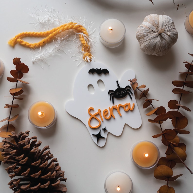 May include: White ghost-shaped wooden ornament with the name "Sierra" written in orange letters. The ornament has a black bat and spider on it and is decorated with glitter.