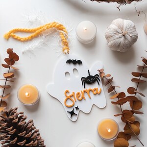 May include: White ghost-shaped ornament with a black spider and bat, orange lettering that says "Sierra", and a yellow yarn hanger.