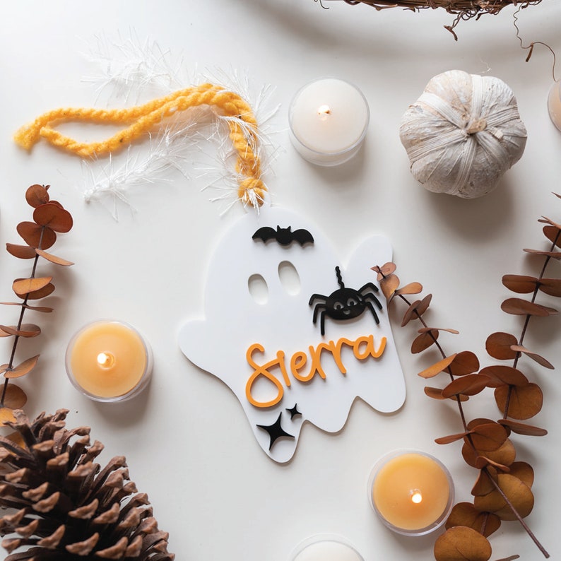 May include: White ghost-shaped ornament with a black spider and bat, orange lettering that says "Sierra", and a yellow yarn hanger.
