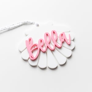 May include: A white seashell-shaped ornament with the name "Bella" written in pink lettering.