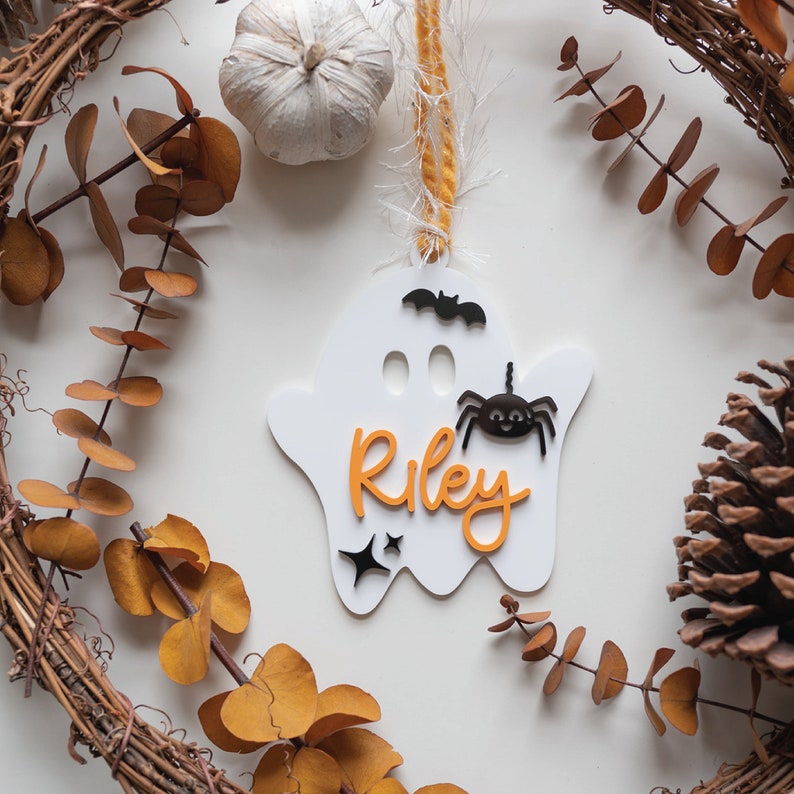 May include: White ghost-shaped ornament with the name "Riley" in orange letters, a black spider, and a black bat. The ornament is hanging from a white string with a yellow and white tassel.