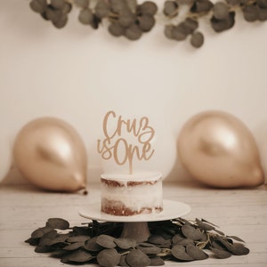 Puede incluir: Un pastel peque&ntilde;o y redondo con glaseado blanco y un adorno de pastel de madera que dice "Cruz is One". El pastel est&aacute; sobre un plato de pedestal blanco. El fondo incluye globos dorados y blancos y vegetaci&oacute;n.