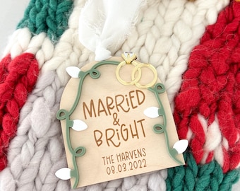 May include: A wooden Christmas ornament shaped like a house with a green garland and white lights. The ornament says "Married & Bright" with the date "08.03.2022" and the names "The Harvens".