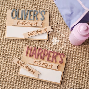 May include: Two wooden signs with the names "Oliver's" and "Harper's" in gray and pink, respectively. Each sign has a white rectangular space for writing and banners that read "first day of" and "last day of". A pink water bottle and a blue bag are also visible.