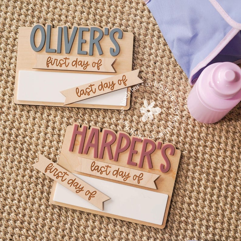 May include: Two wooden signs with the names "Oliver's" and "Harper's" in gray and pink, respectively. Each sign has a white rectangular space for writing and banners that read "first day of" and "last day of". A pink water bottle and a blue bag are also visible.