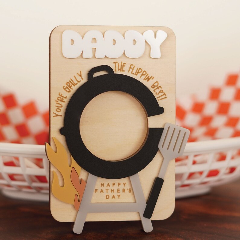 May include: Wooden Father's Day card with a barbecue design. The card says "Daddy, You're Grilly the Flippin' Best!" and "Happy Father's Day".