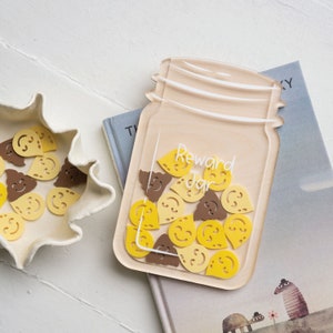 May include: A clear acrylic jar-shaped container labelled "Reward Jar" filled with yellow and brown smiley face tokens. The jar sits on a book with a whimsical illustration. A white, ruffled dish holds additional tokens.