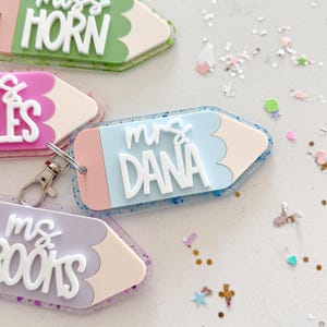 Pode incluir: Quatro chaveiros em forma de l&aacute;pis coloridos com detalhes de glitter. Cada chaveiro tem o nome de um professor diferente: "Miss Horn", "Mrs. Dana", "Mrs. Books" e "Mrs. Giles".