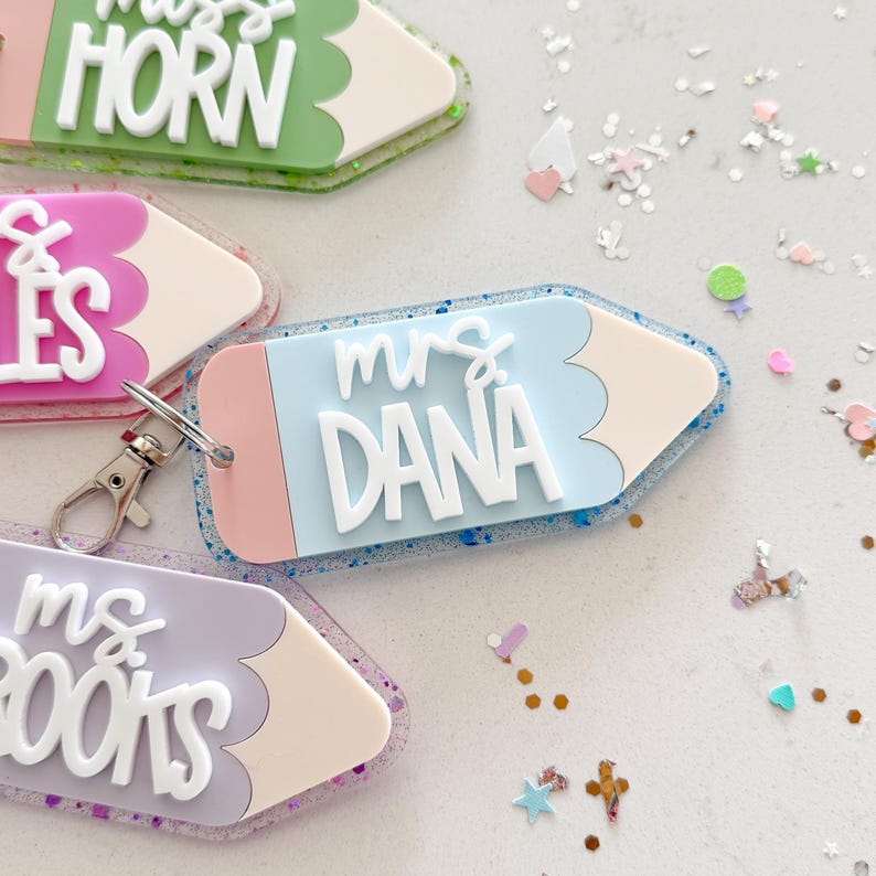 Pode incluir: Quatro chaveiros em forma de l&aacute;pis coloridos com detalhes de glitter. Cada chaveiro tem o nome de um professor diferente: "Miss Horn", "Mrs. Dana", "Mrs. Books" e "Mrs. Giles".