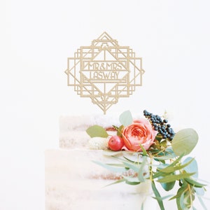 May include: A wooden cake topper with an art deco design. The topper features the text "MR & MRS LASWAY" in a geometric frame.