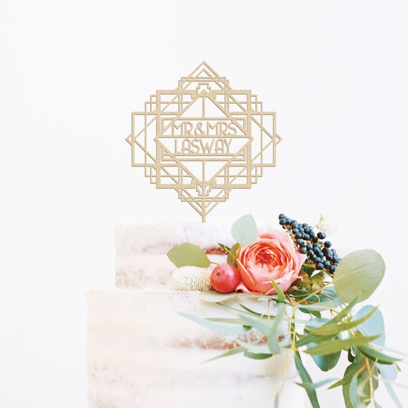 May include: A wooden cake topper with an art deco design. The topper features the text "MR & MRS LASWAY" in a geometric frame.