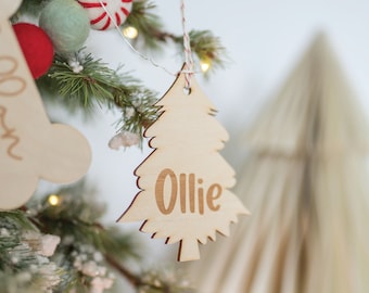 Könnte beinhalten: Ein hölzerner Weihnachtsbaumschmuck mit dem Namen "Ollie" eingraviert. Der Schmuck hängt an einem Weihnachtsbaum mit anderem Schmuck.