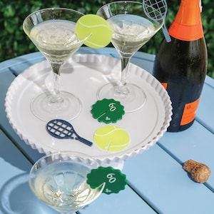 Peut inclure: Deux verres à martini remplis d'un liquide clair, garnis de décorations en forme de balle de tennis, sont posés sur un plateau blanc. Une bouteille de champagne et des marqueurs de boissons sur le thème du tennis sont également présents.