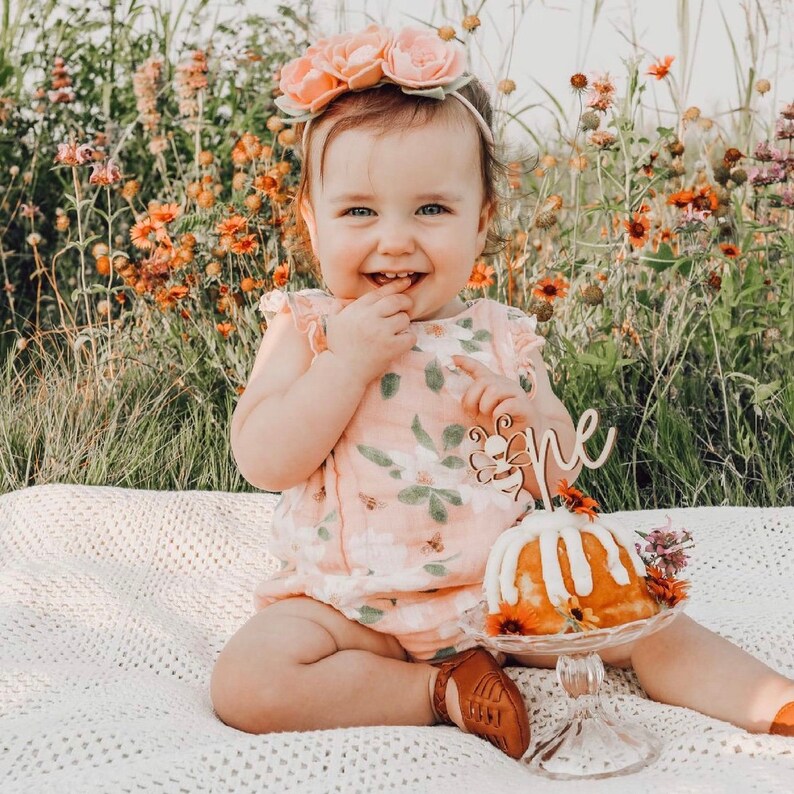 May include: A smiling baby wearing a floral romper and a flower crown sits on a white blanket in a field of wildflowers. A small cake with a wooden "one" cake topper sits in front of the baby.