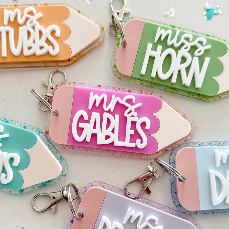 Pode incluir: Cinco chaveiros em forma de l&aacute;pis coloridos com detalhes de glitter. Cada chaveiro tem o nome de um professor diferente: "Mrs. Tubbs", "Miss Horn", "Mrs. Gables", "Mrs. Davis" e "Mrs. DePoy".