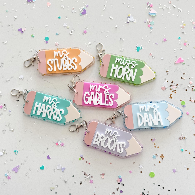 Pode incluir: Seis chaveiros em forma de l&aacute;pis coloridos com diferentes nomes de professores. Os chaveiros s&atilde;o feitos de acr&iacute;lico transparente com glitter embutido no pl&aacute;stico. Os nomes nos chaveiros s&atilde;o "Ms. Stubbs", "Miss Horn", "Mrs. Gables", "Ms. Harris", "Mrs. Dana" e "Ms. Brooks".