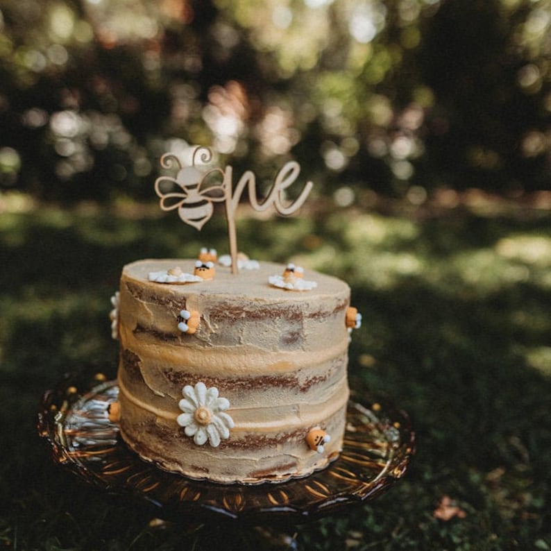 May include: A small, round cake with tan frosting and decorative elements. The cake is adorned with small bee and flower decorations, and a wooden topper that reads "one" with a bee design. The cake sits on a decorative glass plate, with a blurred green background.