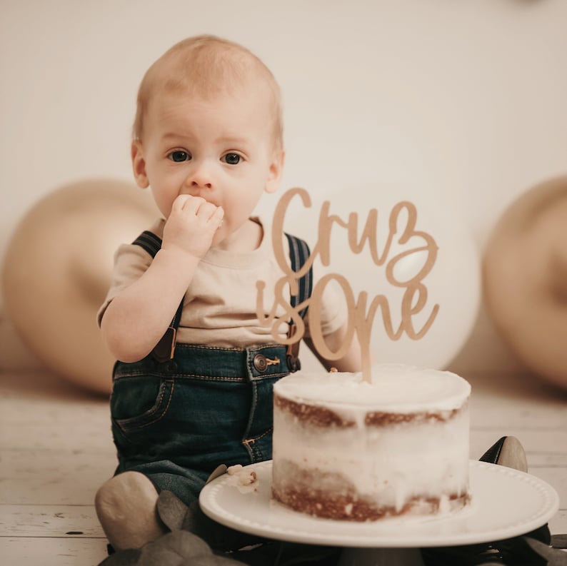 Puede incluir: Un ni&ntilde;o peque&ntilde;o con un peto vaquero y una camiseta color beige est&aacute; sentado cerca de un pastel peque&ntilde;o con una decoraci&oacute;n de madera que dice "Cruz is One". El pastel est&aacute; sobre un plato blanco, con globos dorados y blancos en el fondo.