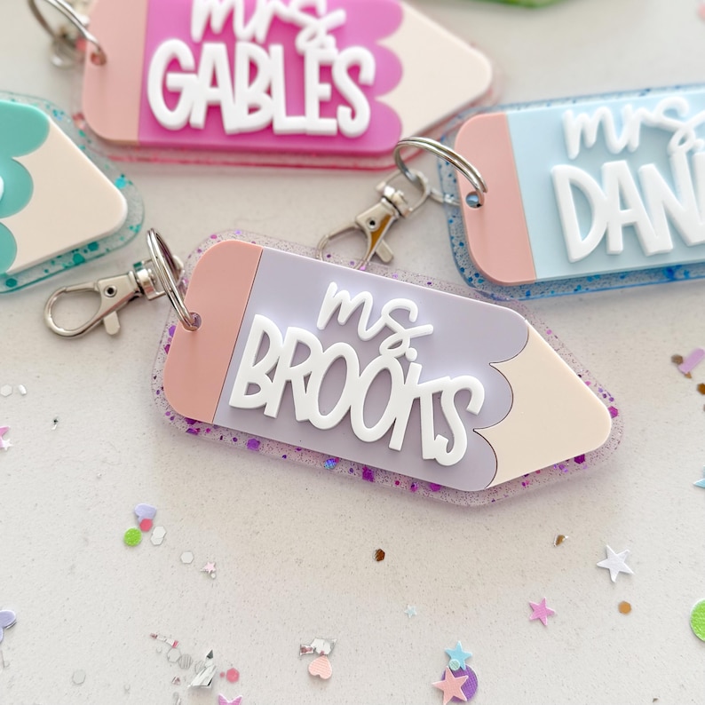 Pode incluir: Um chaveiro em forma de l&aacute;pis roxo e rosa com detalhes de glitter e o texto "ms. brooks" em letras brancas em relevo.
