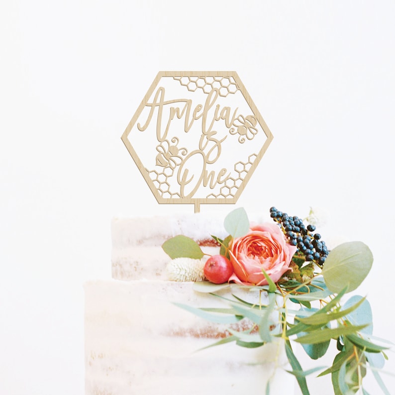 May include: A wooden cake topper in the shape of a hexagon with a honeycomb pattern and the text "Amelia is One" with bees. The topper is on a white cake with pink and green flowers.