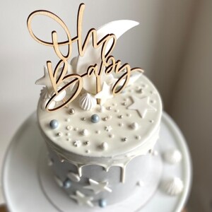 May include: A white cake with gray frosting and a wooden "Oh Baby" cake topper with a crescent moon and stars. The cake is decorated with white and blue sprinkles and stars.