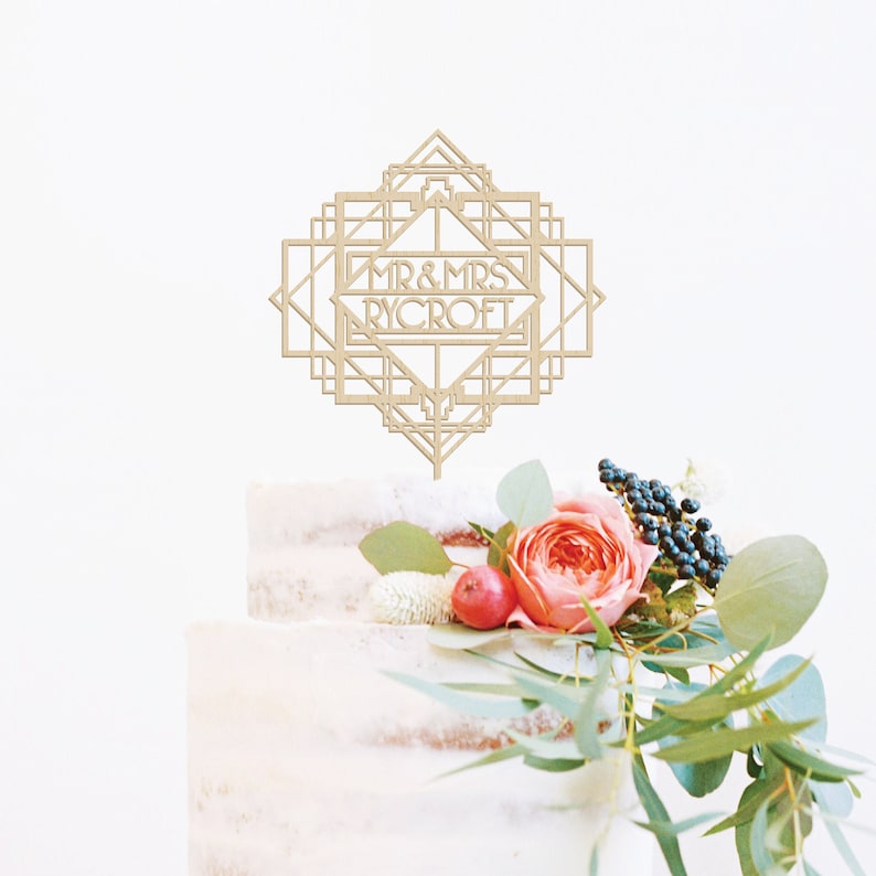 May include: A wooden cake topper with an art deco design and the text "MR & MRS RYCROFT". The topper is on a white cake with pink and green flowers.