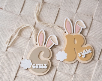 May include: Two wooden bunny-shaped ornaments with the names "Cruz" and "Rylie" written on them. The ornaments are beige and yellow with pink bunny ears and white fluffy tails.