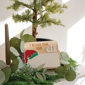 Könnte beinhalten: Ein Holzschild mit dem Text "A message from your elf" und einer Grafik eines roten und grünen Elfenhutes. Das Schild hat einen leeren weißen Bereich zum Schreiben einer Nachricht.