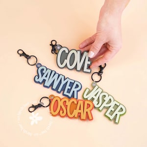 May include: Keyrings with names: "COVE", "SAWYER", "OSCAR", and "JASPER". Each keyring features a name in bold, layered lettering with a carabiner clip. The names are in various colours: grey, blue, orange, and green. A hand is holding one of the keyrings.