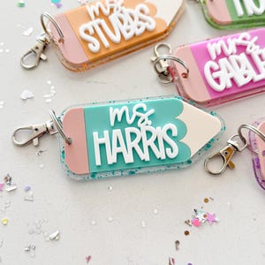 Pode incluir: Um chaveiro em forma de l&aacute;pis azul turquesa e rosa com detalhes de glitter. O chaveiro tem o texto "ms. HARRIS" em letras brancas.