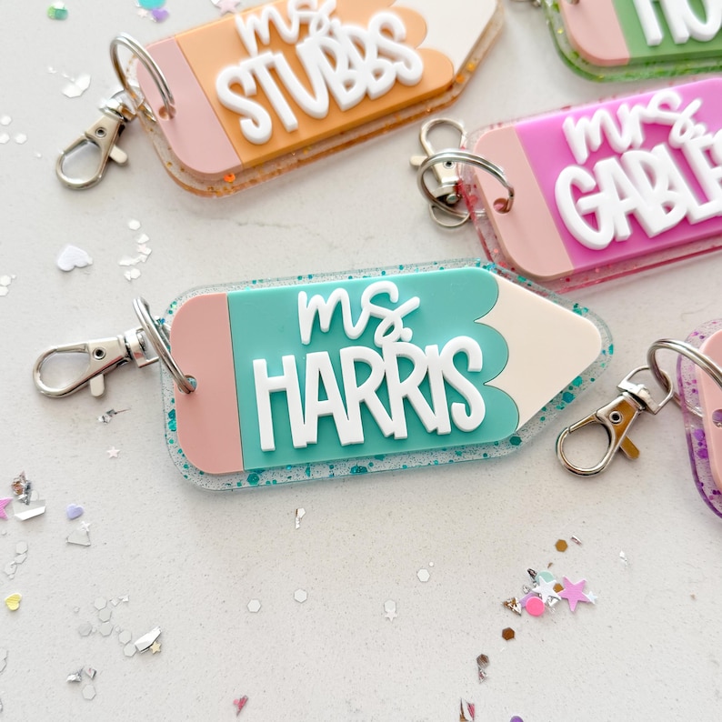 Pode incluir: Um chaveiro em forma de l&aacute;pis azul turquesa e rosa com detalhes de glitter. O chaveiro tem o texto "ms. HARRIS" em letras brancas.