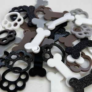 May include: Assortment of small, colourful, plastic paw print and bone shapes. The shapes are in various colours, including black, white, brown, and grey. Some of the shapes have glitter.