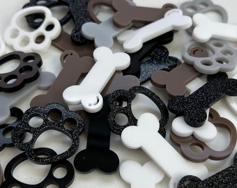 May include: Assortment of small, colorful, plastic paw print and bone shapes. The shapes are in various colors, including black, white, brown, and gray. Some of the shapes have glitter.