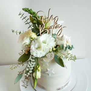 May include: A white cake with a wooden "Oh Baby" topper and white flowers. The cake is decorated with white stars.