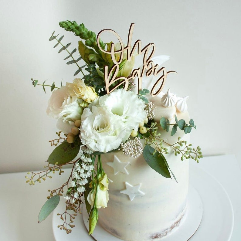 May include: A white cake with a wooden "Oh Baby" topper and white flowers. The cake is decorated with white stars.