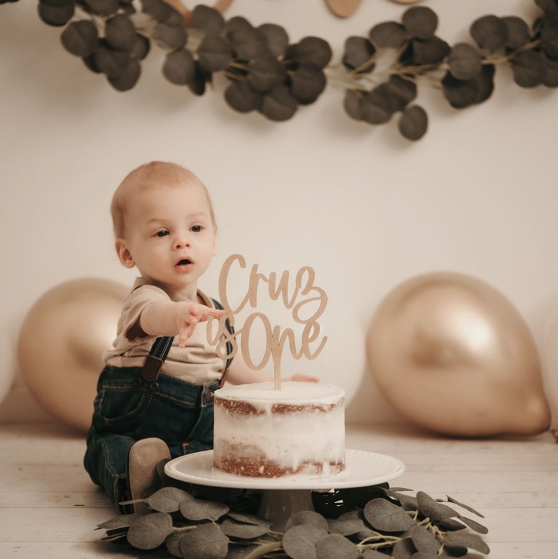 Puede incluir: Una peque&ntilde;a tarta glaseada con un adorno de pastel de madera que dice "Cruz is One". La tarta est&aacute; sobre un pedestal blanco, con un beb&eacute; con peto vaquero y tirantes se&ntilde;alando la tarta. Globos dorados y blancos en el fondo.