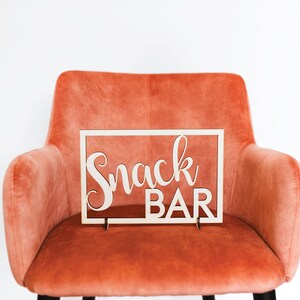Puede incluir: Un letrero de madera con las palabras "Snack Bar" en una fuente cursiva, sentado en un sill&oacute;n de terciopelo coral.
