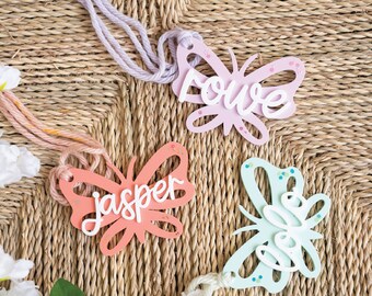 May include: Three butterfly-shaped ornaments with names written on them. The butterflies are pink, orange, and light blue with white lettering. The names are "Love", "Jasper", and "Elle".