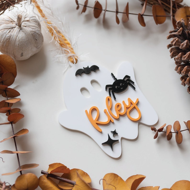May include: White ghost-shaped ornament with the name Riley in orange letters, a black spider, and a black bat. The ornament has a gold string for hanging.